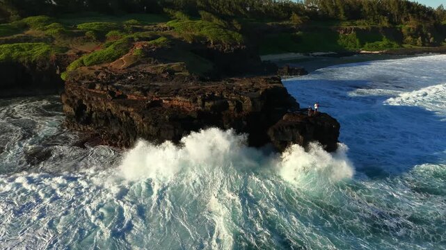 Black volcanic rock juts into the ocean. Waves crash against its rugged base. Bright blue water reflects sunlight. Lush green hills form the background. Captures raw coastal beauty
