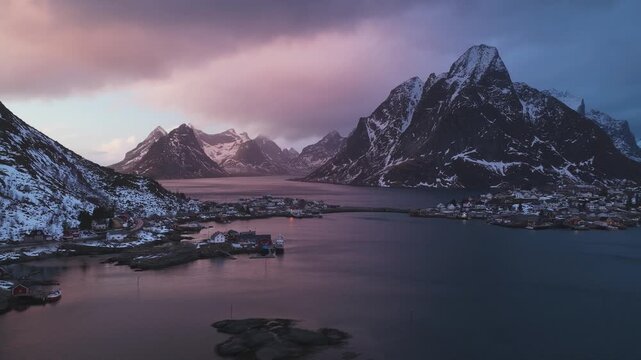 Aerial view of Reinebringen's majestic snow-dusted peaks and the charming village nestled along the water's edge, bathed in the soft glow, Reine, Nordland, Norway.