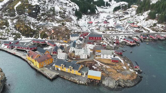 Aerial view of colorful buildings nestled between the sea and snow-dusted mountains in the scenic Nusfjord fishing village, Ramberg, Nordland, Norway.