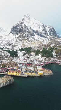 Aerial view of the colorful Nusfjord fishing village nestled against snow-capped mountains with the teal waters of the Norwegian Sea, Nusfjord, Nordland, Norway.