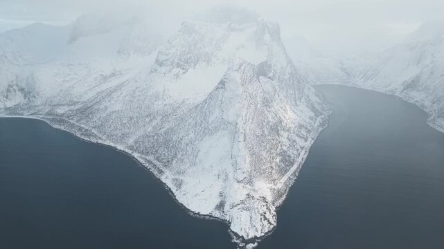Aerial view of the snow-covered Segla mountain peak meeting the dark fjord waters, showcasing the stark contrast, Segla, Troms, Norway.