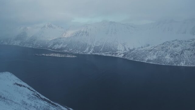 Aerial view of Segla mountain's snow-capped peaks tower above the dark fjord waters, a stark contrast against the cloudy sky, Segla, Troms, Norway.