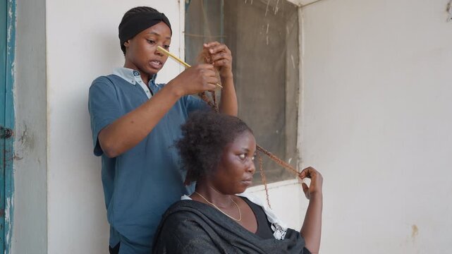 Black stylist braiding client hair, standing apprentice combing textured curls on seated client, focused expression, hands working with comb, casual doorway setting, collaborative grooming scene,
