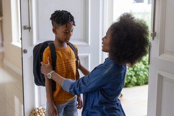 Fototapeta premium Diverse mother and child at door, mother kneeling, adjusting backpack, child wearing orange tee