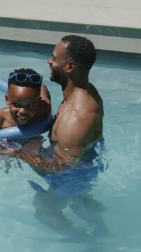 Vertical video: Child giggling as father in trunks steadying child in pool with ring, copy space