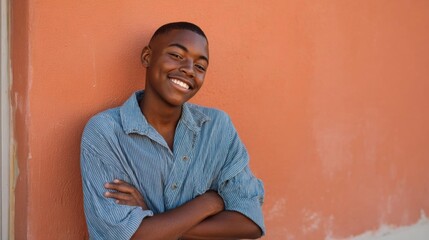 Obraz premium Portrait of a young african-american man standing in front of an orange wall. he is wearing a blue striped button-down shirt and has his arms crossed over his chest.