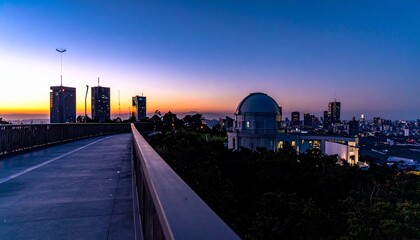 Fototapeta premium City skyline at sunset with dome building, pedestrian bridge leading forward, skyscrapers behind, sky blending deep blue to warm orange.