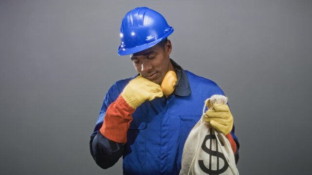 Construction worker man in blue hardhat and coveralls holding a dollar sack and touching earmuff with hand on chin pensive pose in studio; pondering.