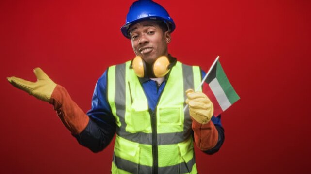 Black construction worker in blue hardhat wearing high visibility vest and ear protectors holds kuwait flag and shrugs with gloved hands in studio; pride solidarity.