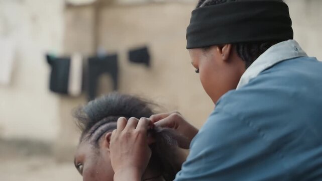 Outdoor closeup braiding hair gently. Home stylist in beanie carefully parting scalp while plaiting tight rows, hands focused, neighbor seated, laundry on wall in background, soft daylight, intimate