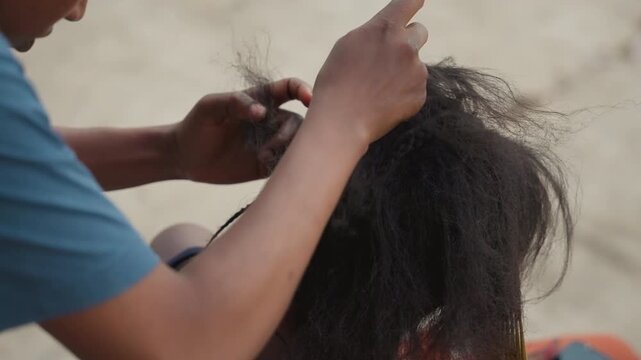 Hands detangling thick natural hair. Mobile braider loosening tight tangles with fingertips, careful pulling to prevent breakage, sandy ground, seated client, rhythm of treatment, resilient texture,