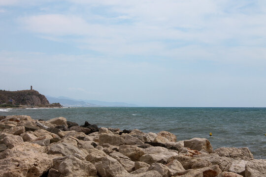 A serene wide shot of the rugged Mediterranean shoreline near Torre del Mar, Spain. In the foreground, weathered rocks and stones are scattered in the clear turquoise water, while the light blue sky
