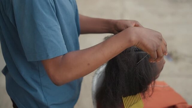 Stylist parting hair and braiding outdoors with comb and steady hands, visible scalp rows and protective styling on bench, natural light, rhythmic technique, mobile salon atmosphere and close
