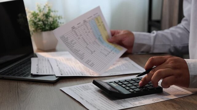 Close-up of a person calculating numbers with a calculator while filling out documents at a desk with a laptop in a home or office setting, suitable for finance, accounting, and paperwork tasks.