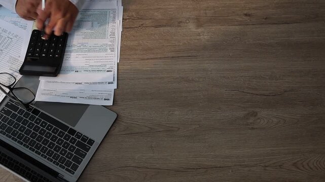 Top view of a person reviewing budget planning documents with a calculator on a desk next to a laptop and glasses, with copy space on a wooden table, suitable for finance, tax filing, and accounting c