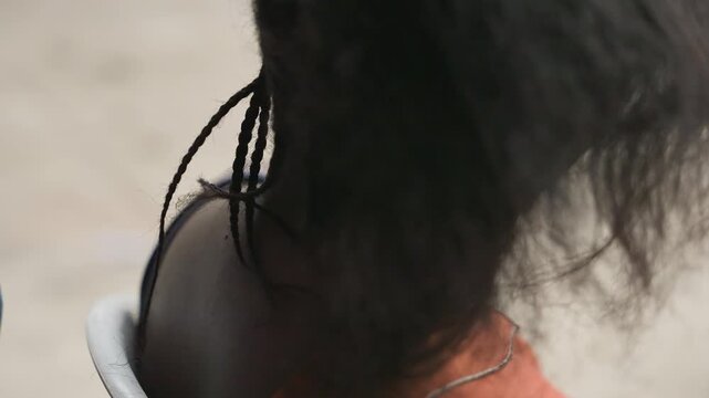 Closeup hair braid at nape, hands pulling and tightening small plait at back of head, soft outdoor light, white chair and orange top visible, textured natural hair, patient fingertip work, intimate