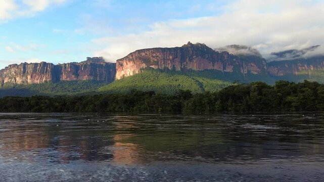 POV traveling along the Churun River, with calm waters and lush vegetation, a giant tepui in the background, illuminated by the sun's rays, and orange-colored rock, Canaima National Park, Venezuela