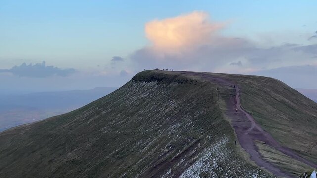 Pen y Fan summit ridge with snow patches and hikers walking along trail under sunset sky creating dramatic mountain landscape with depth and scenic atmosphere