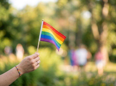 Close-up of a hand holding a rainbow pride flag in a sunny outdoor setting, symbolizing LGBTQ+ pride, inclusion, and equality.