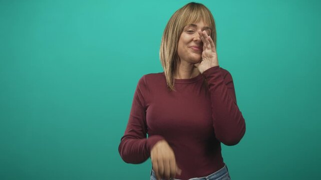 Woman cupping hands around mouth shouting in studio while smiling, maroon longsleeve and jeans visible; joyful announcement.