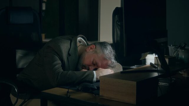 Medium shot of exhausted mature Caucasian man in suit jacket leaning on folded hands and sleeping with head on desk, while working to deadline on computer in darkness alone in office at night
