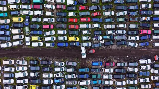 A large collection of cars fills a lot with many colors and styles. The vehicles are parked closely together with a dirt path running through the middle. It is daytime and bright.