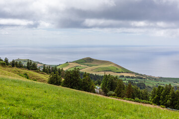Fototapeta premium Atlantic View, San Miguel Island, Azores, Portugal. Rolling hills and green fields under a cloudy sky. Distant ocean visible in the background. Rural landscape with scattered trees.