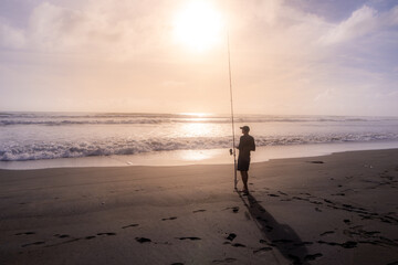 A lone fisherman stands silhouetted against the sun, casting his line into the ocean at Karekare Beach, Auckland, New Zealand He is hoping to catch fish. © Zenstratus