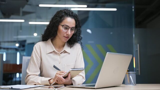 Businesswoman watching video call online conference taking notes looking at laptop computer screen sitting at workplace in office. Female employee listening remote business training, course or seminar