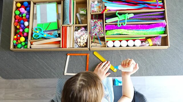 Top view of little girl making craft from wooden sticks at table using colorful materials. Kids creativity fine motor skills development and hands on learning concept.