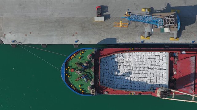 Aerial view of a red cargo ship filled with white packages near a concrete dock with blue cranes, contrasting against the teal water, Ortona, Abruzzo, Italy.