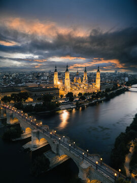 Aerial view of the illuminated Basilica of Our Lady of the Pillar and the stone bridge over the Ebro river at sunset in Zaragoza, Aragon, Spain.