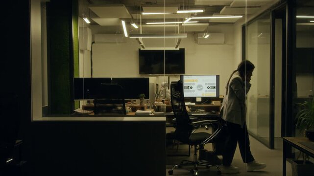 Wide shot through office window of young female corporate employee sitting at computer with feet up in office while working at midnight, arguing on phone with husband while pacing around