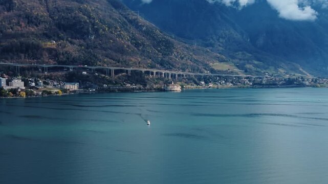 Aerial view of a boat sailing on the clear water near Chillon Castle under a bridge, set against a backdrop of majestic mountains, Montreux, Vaud, Switzerland.