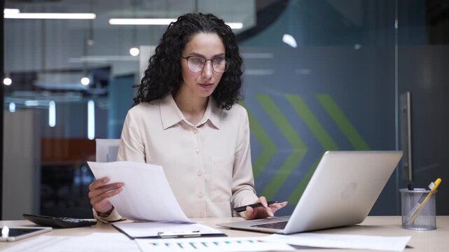 Happy businesswoman satisfied with financial results sitting at workplace in office. Smiling female financier looks at documents and computer, rejoices at positive indicators, celebrates success