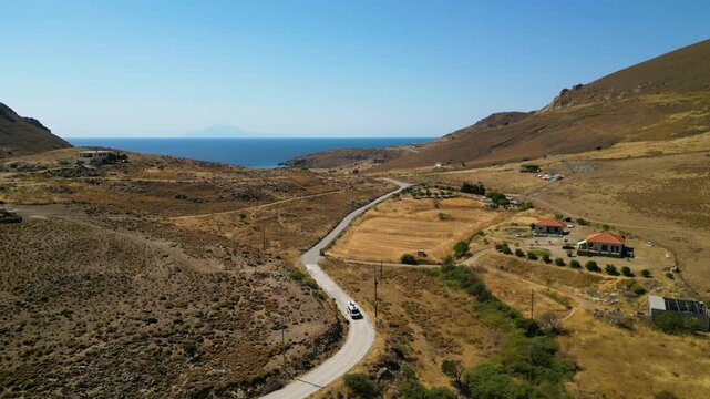 Aerial view of a white camper van traveling along a winding dirt road through the rugged, golden landscape of Limnos Island, Greece, highlighting the freedom of off-grid exploration.