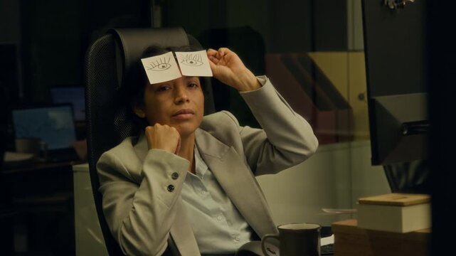 Medium portrait of exhausted, weary, apathetic Hispanic female manager working on project on computer at midnight in office, snoozing with cartoon eyes on glasses, taking off and looking at camera