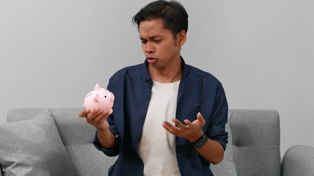 Unhappy asian man at home with empty piggy bank with a few coins inside, feeling disappointed, difficult situation. Bankruptcy