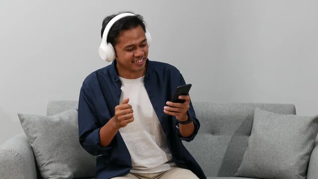 Happy carefree Young Asian man in stylish headphones listening to music from smartphone moving head, hand to rhythm