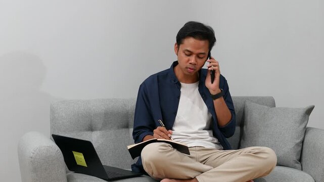 Young Asian man writing notes sitting on couch at home, Ordering goods or communicating with colleagues