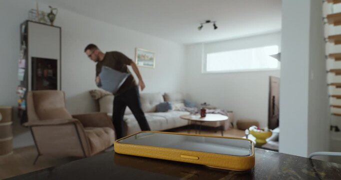 Focus on a smartphone left on a table while a man searches around the living room in the background, showing forgetfulness, misplaced device, and everyday home routine in a realistic domestic setting.
