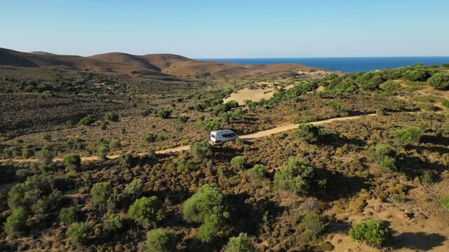 Aerial view of a white camper van traveling along a winding dirt road through the rugged, golden landscape of Limnos Island, Greece, highlighting the freedom of off-grid exploration.