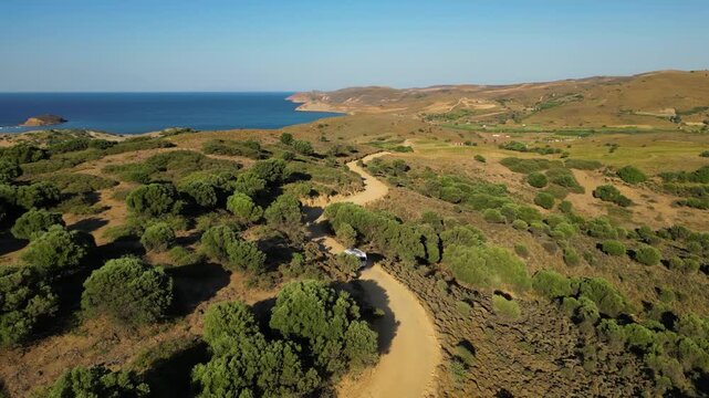 Aerial view of a white camper van traveling along a winding dirt road through the rugged, golden landscape of Limnos Island, Greece, highlighting the freedom of off-grid exploration.