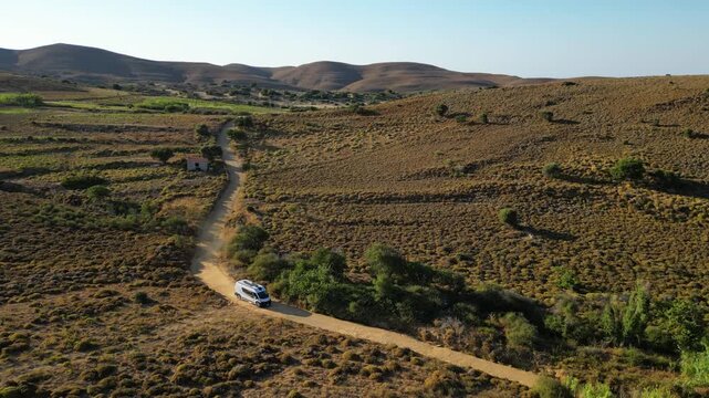 Aerial view of a white camper van traveling along a winding dirt road through the rugged, golden landscape of Limnos Island, Greece, highlighting the freedom of off-grid exploration.