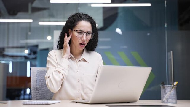 Sick businesswoman suffers from ear pain while sitting at workplace in office. Upset woman touches her ear with her fingers. She has furuncle, shingles, eczema, ear diseases or otitis media. Earache