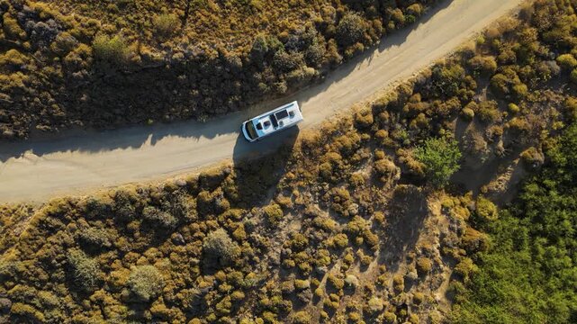 Aerial view of a white camper van traveling along a winding dirt road through the rugged, golden landscape of Limnos Island, Greece, highlighting the freedom of off-grid exploration.