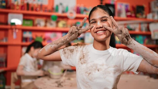 Happy caucasian girl pose at camera while diverse children modeling clay behind. Cute student wearing dirty shirt while looking at camera at workshop in art lesson. Blurring background. Edification