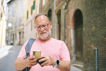 Senior man using smartphone while walking on quiet old street, wearing glasses and casual clothes,...