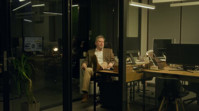 Full shot of Caucasian male manager nodding off at desk with feet up while resting from working on report late at night, waking up, grabbing documents, returning for smartphone, walking out of office