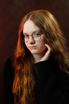 Moody studio portrait of a beautiful teenage girl with long ginger hair wearing round glasses and stylish black sweater on dark studio background.